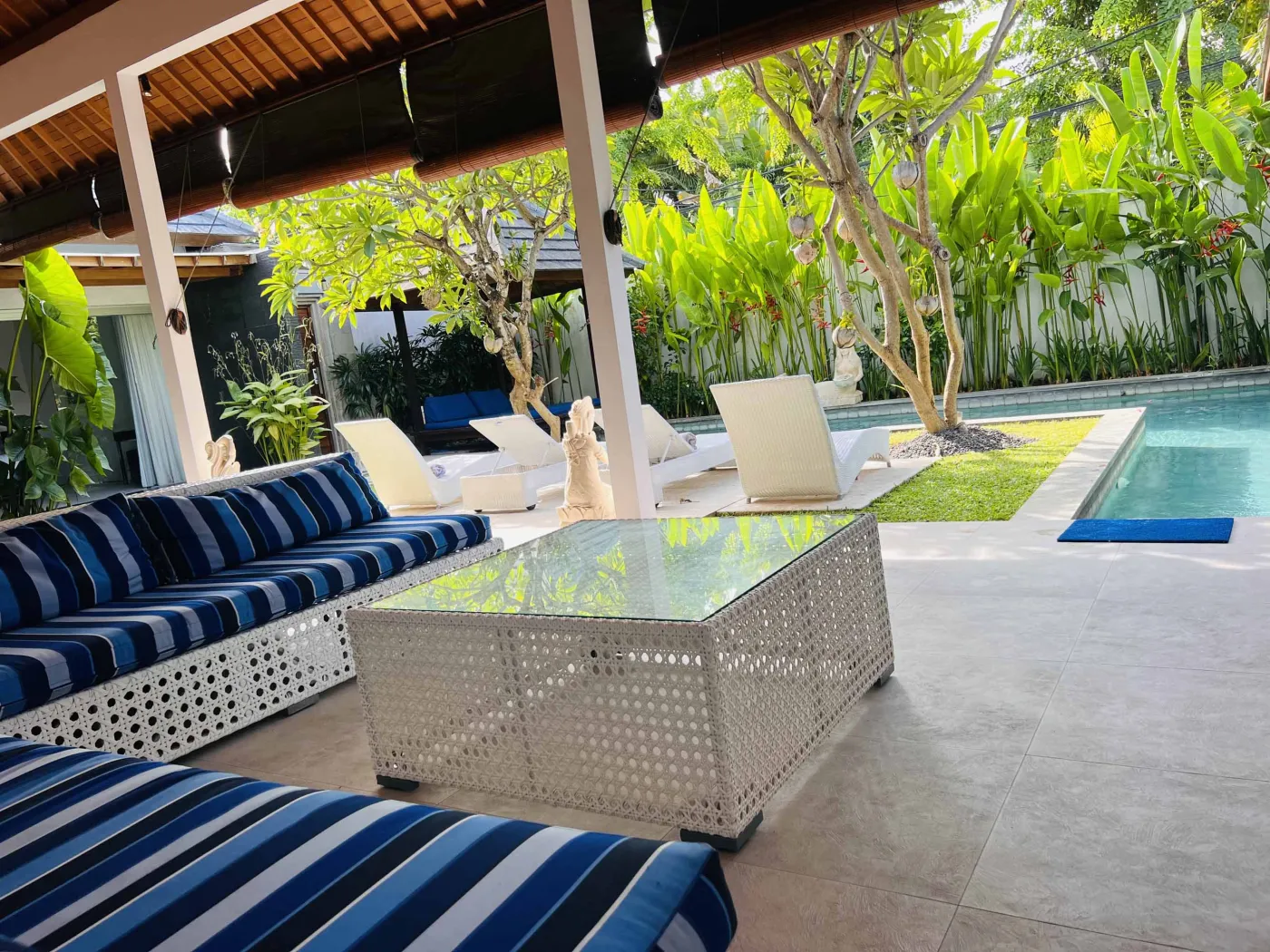 Serene Balinese villa pool with turquoise water, white lounge chairs, frangipani trees, and lush tropical greenery under a blue sky. - Bali Villas
