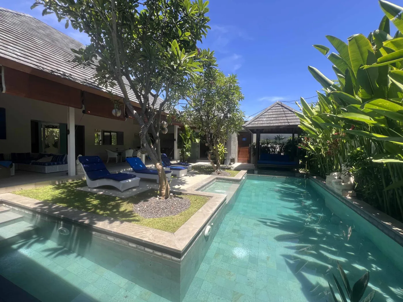 Serene Balinese villa pool with turquoise water, white lounge chairs, frangipani trees, and lush tropical greenery under a blue sky. - Bali Villas