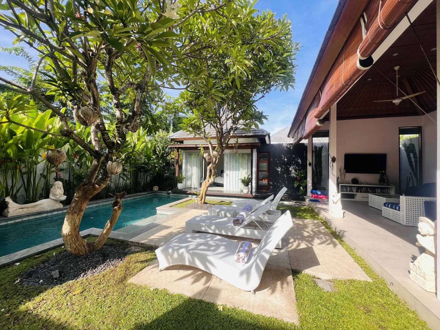 Serene Balinese villa poolside with turquoise water, white lounge chairs, lush tropical greenery, and stone statues under open wooden verandas. - Bali Villas