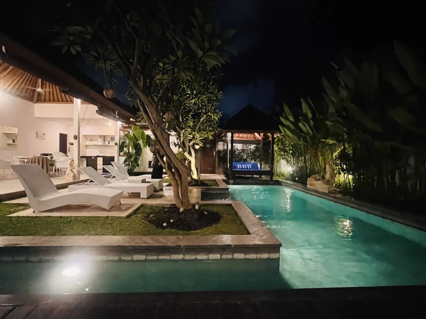 Serene Balinese villa pool with turquoise water, white lounge chairs, frangipani trees, and lush tropical greenery under a blue sky. - Bali Villas