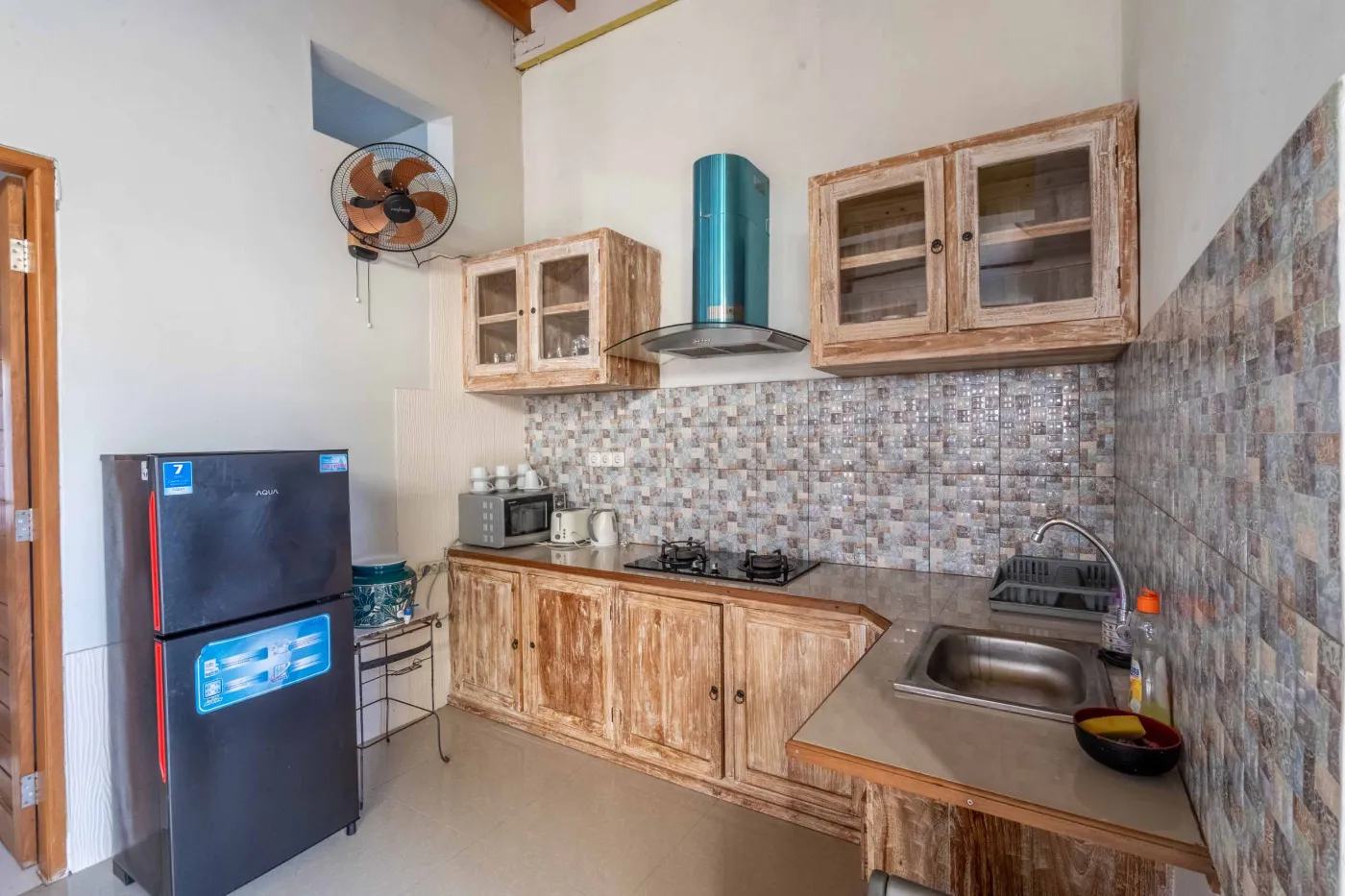 Rustic Balinese kitchen with wooden cabinets, blue range hood, mosaic tiled backsplash, black fridge, and ceiling fan. - Bali Villas