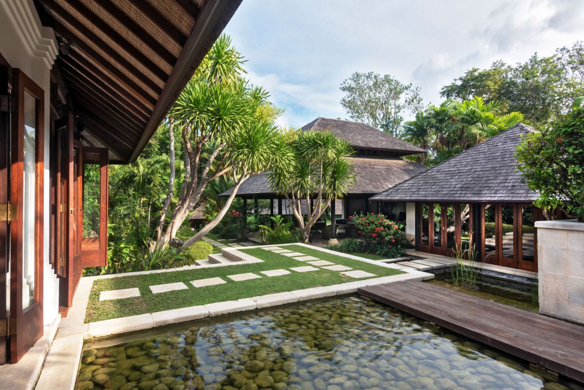 Serene Balinese villa courtyard with koi pond, stone steps, lush tropical plants, and palm trees under a clear sky. - Bali Villas