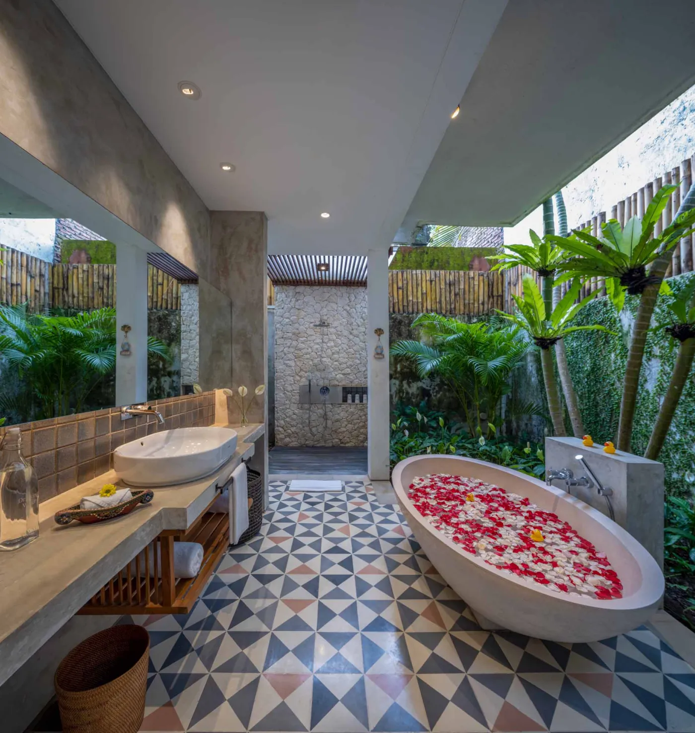 Luxurious open-air Balinese bathroom with freestanding bathtub filled with rose petals, wooden vanity, tropical plants, and geometric tiled floor. - Bali Villas
