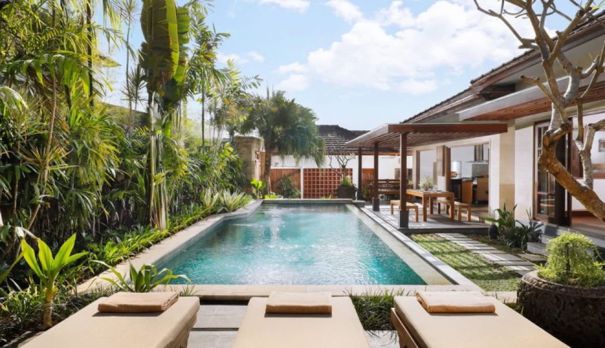 Luxurious Balinese villa infinity pool surrounded by tropical palms, lush greenery, and outdoor seating under blue skies. - Bali Villas