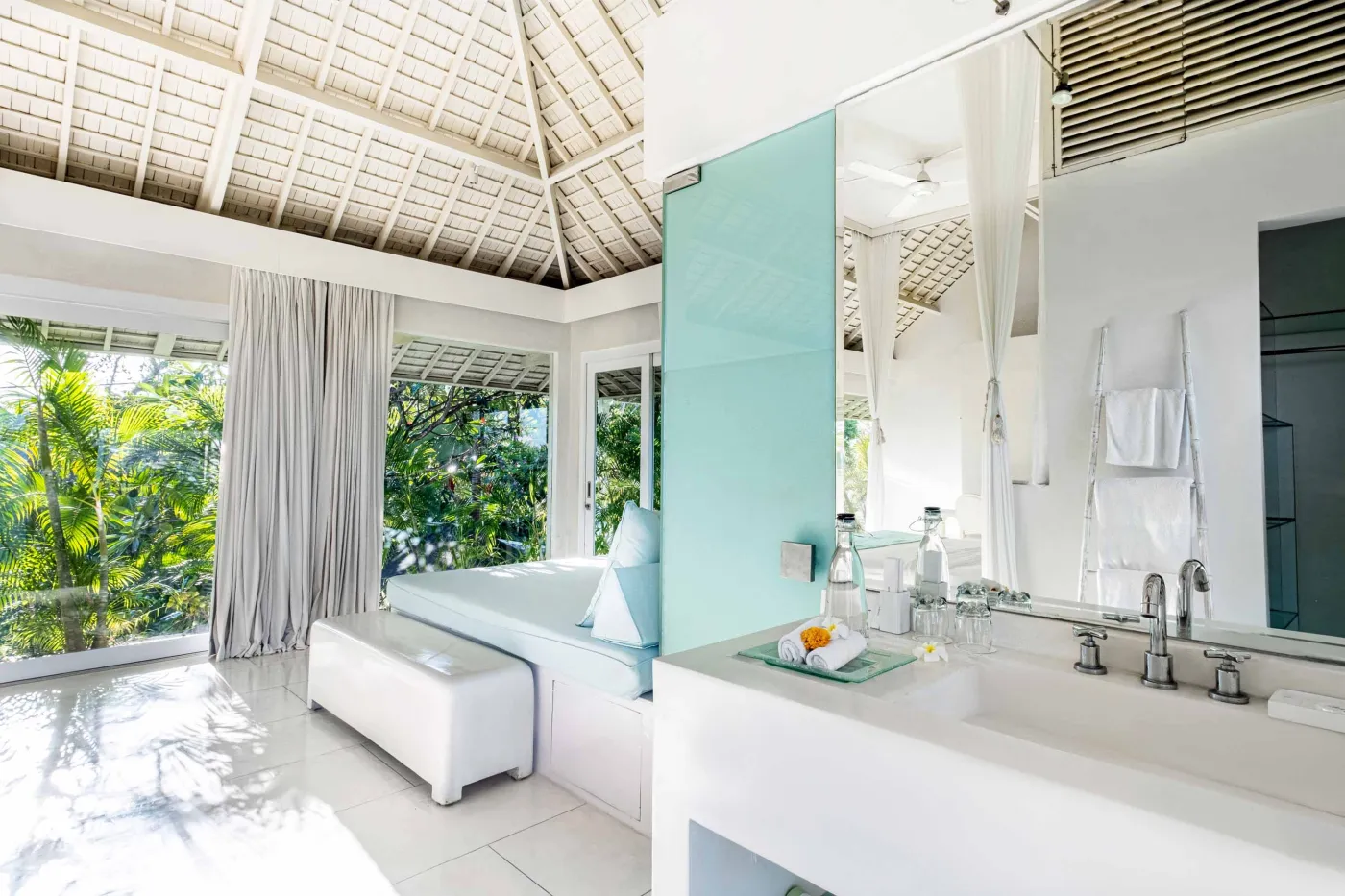 Modern luxury bedroom suite with white platform bed, teal pillow, turquoise sunken bathtub, and garden view through sheer curtains. - Bali Villas