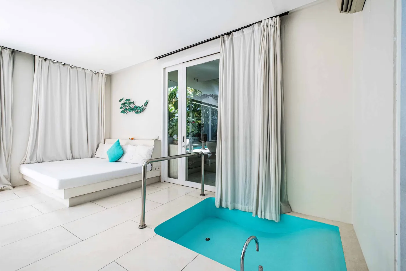 Modern luxury bedroom suite with white platform bed, teal pillow, turquoise sunken bathtub, and garden view through sheer curtains. - Bali Villas