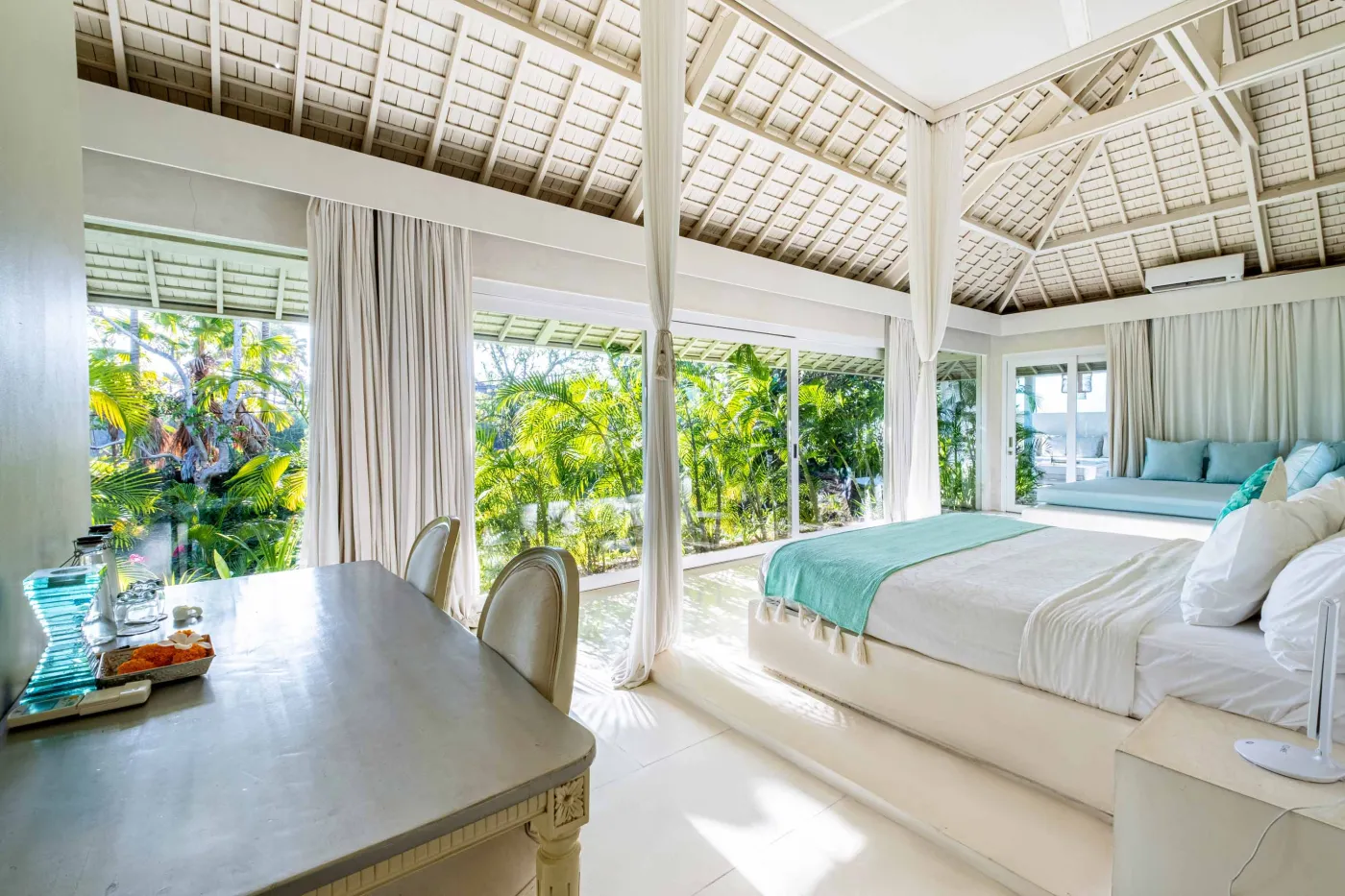 Modern luxury bedroom suite with white platform bed, teal pillow, turquoise sunken bathtub, and garden view through sheer curtains. - Bali Villas