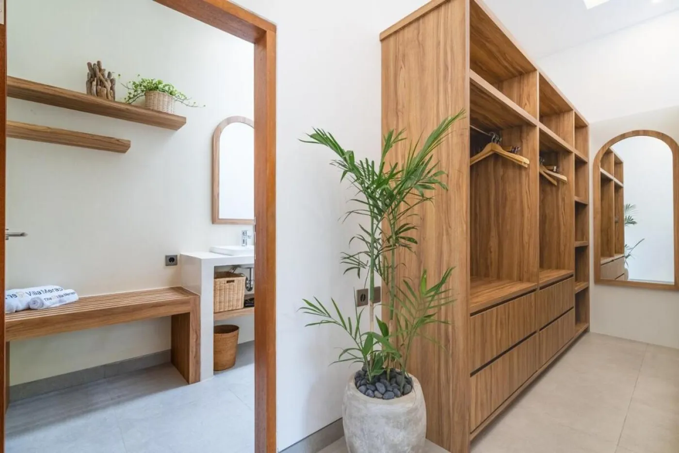 Compact modern bathroom with white toilet, tall potted plant, small shelf of toiletries, and beige walls. - Bali Villas