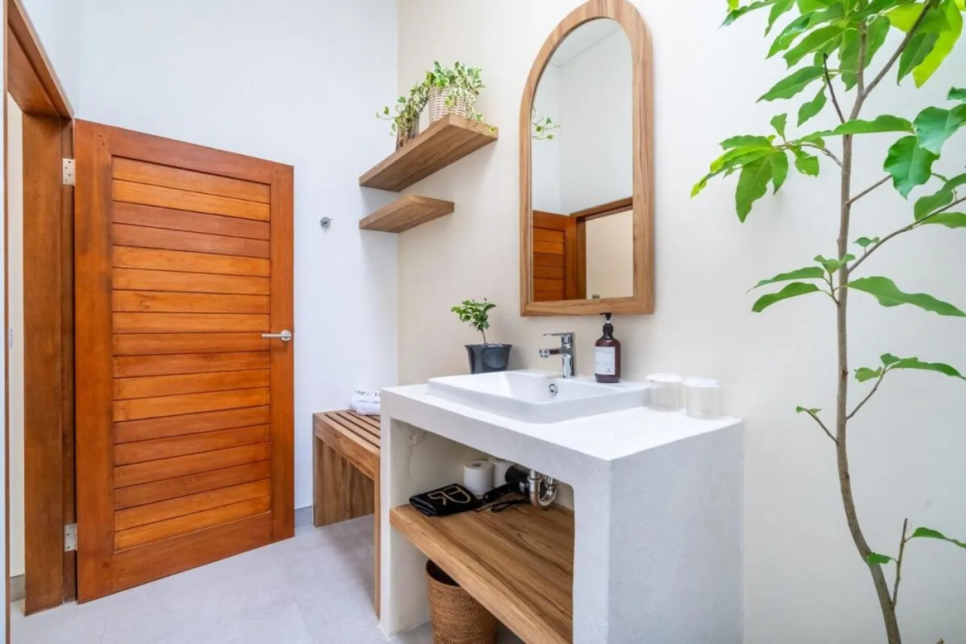 Compact modern bathroom with white toilet, tall potted plant, small shelf of toiletries, and beige walls. - Bali Villas