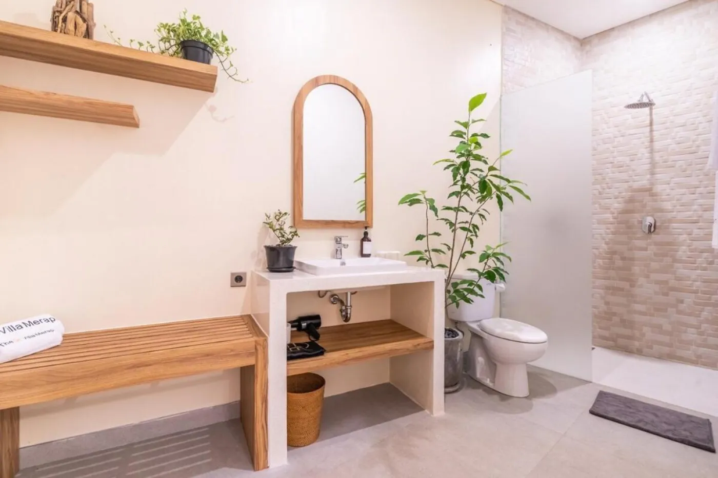 Compact modern bathroom with white toilet, tall potted plant, small shelf of toiletries, and beige walls. - Bali Villas