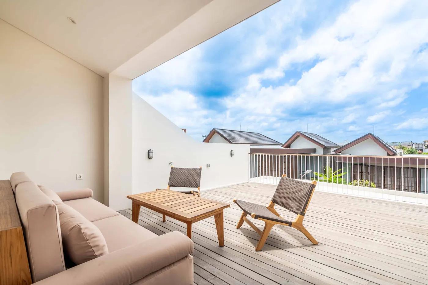 Modern wooden deck balcony with beige sofa, teak table, and chairs, overlooking contemporary rooftops on a sunny day. - Bali Villas