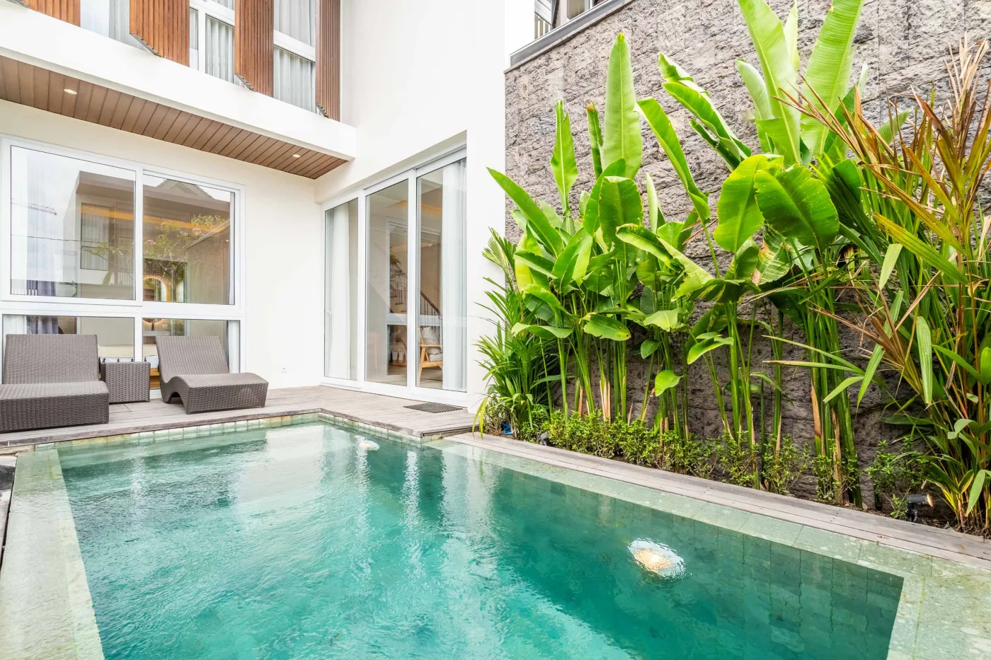 Modern infinity-edge pool in a tropical villa with lounge chairs, lush banana plants, and glass doors to white interior. - Bali Villas