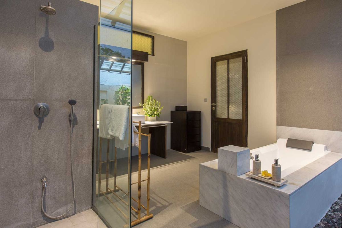 Modern luxury bathroom with freestanding white bathtub, glass shower enclosure, towels, and tropical plant accents. - Bali Villas