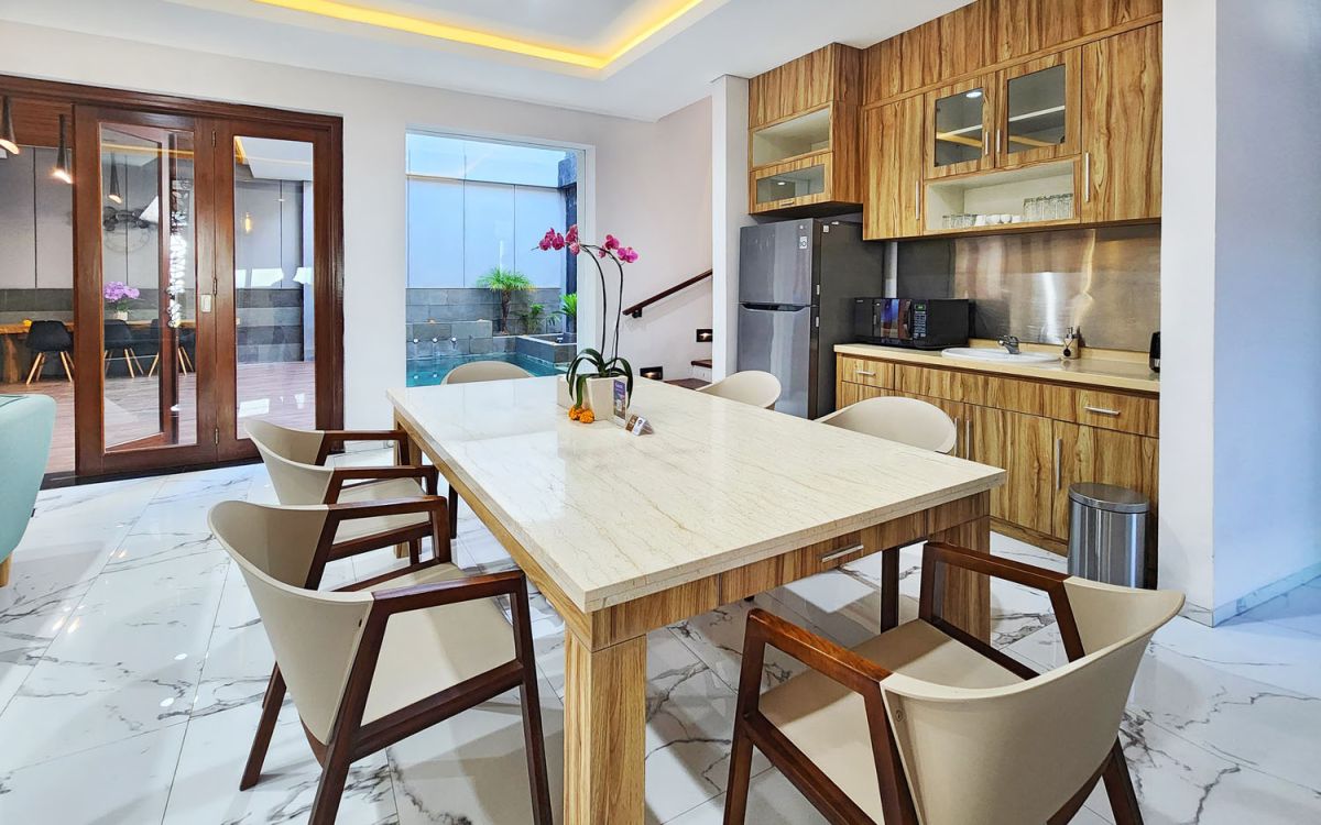 Modern open-plan kitchen and dining area with wooden cabinets, marble-top table, stainless steel fridge, and potted orchids in a Balinese villa. - Bali Villas