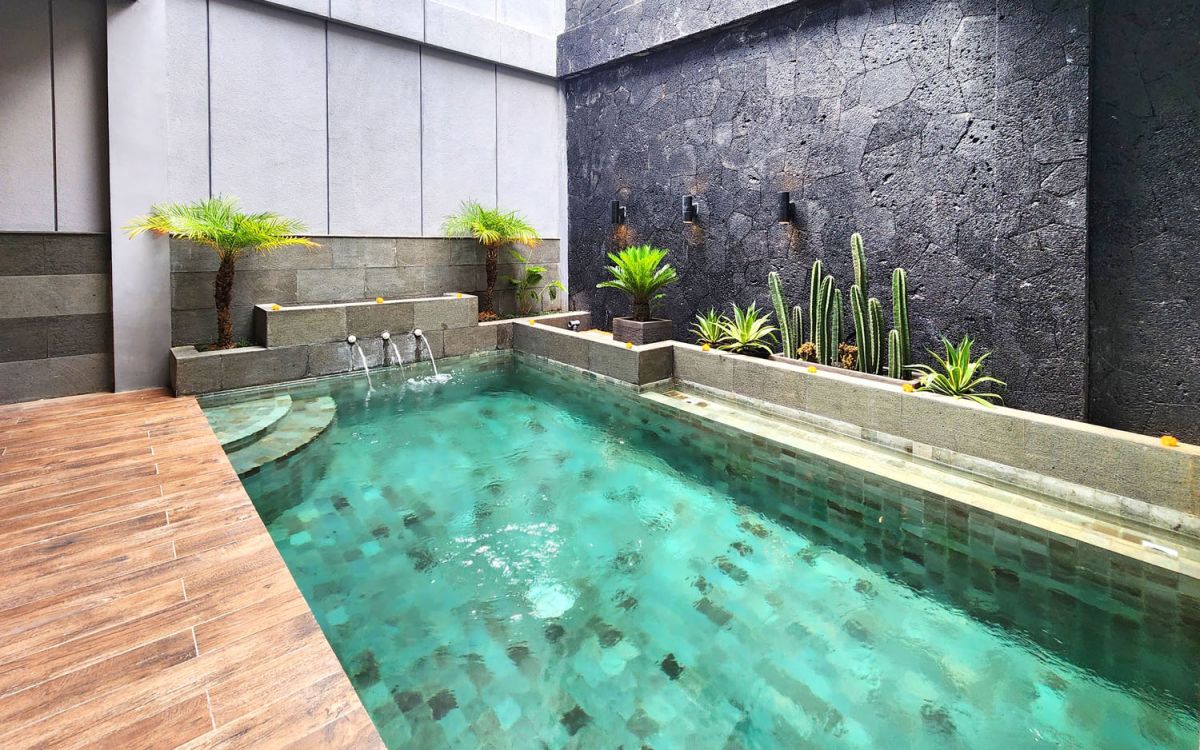 Modern turquoise infinity pool in a tropical courtyard with stone walls, potted palms, cacti, and wooden decking. - Bali Villas