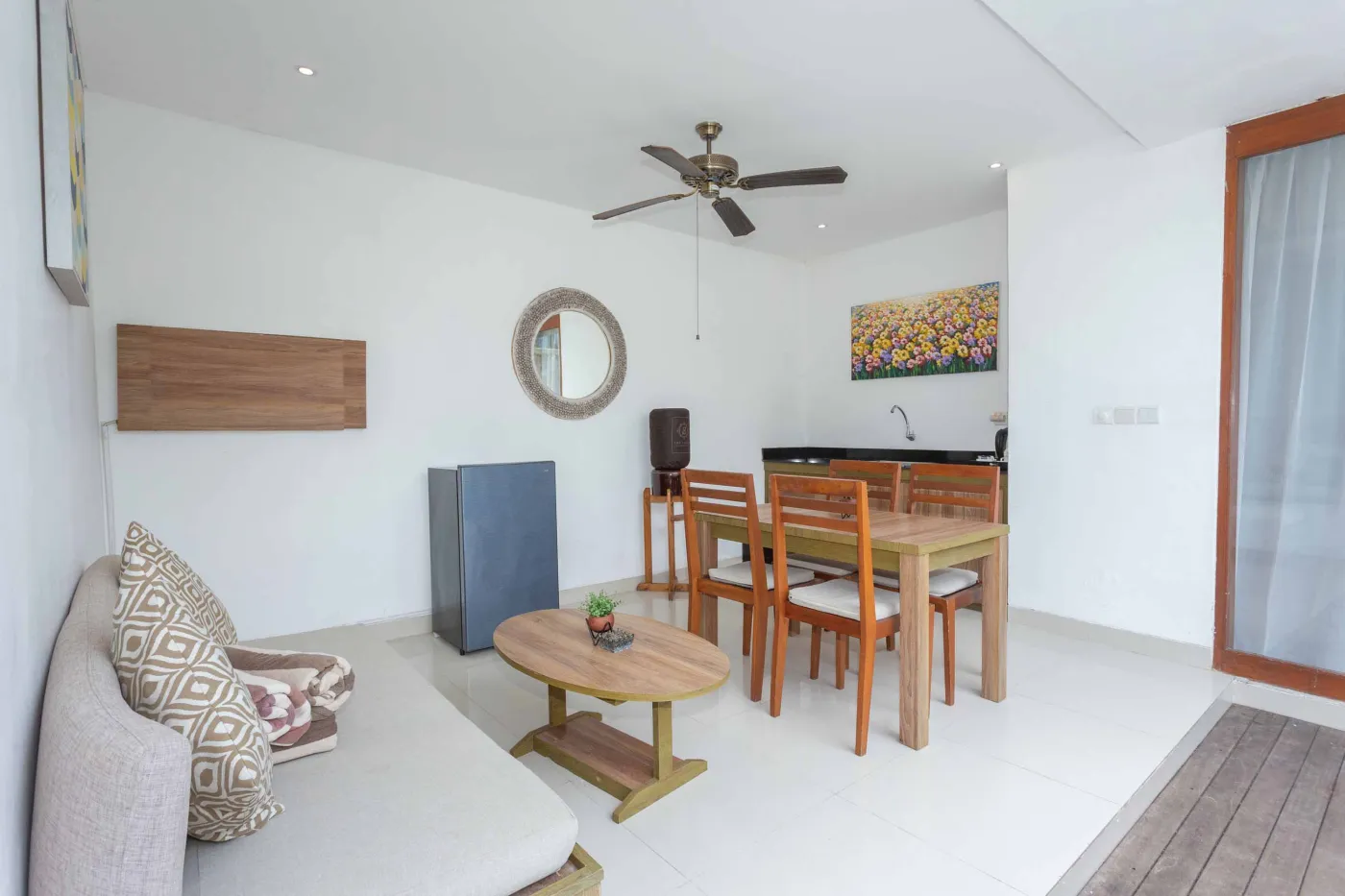 Spacious tropical villa interior with wooden dining table, cushioned sofa, ceiling fan, round mirror, and open deck. - Bali Villas