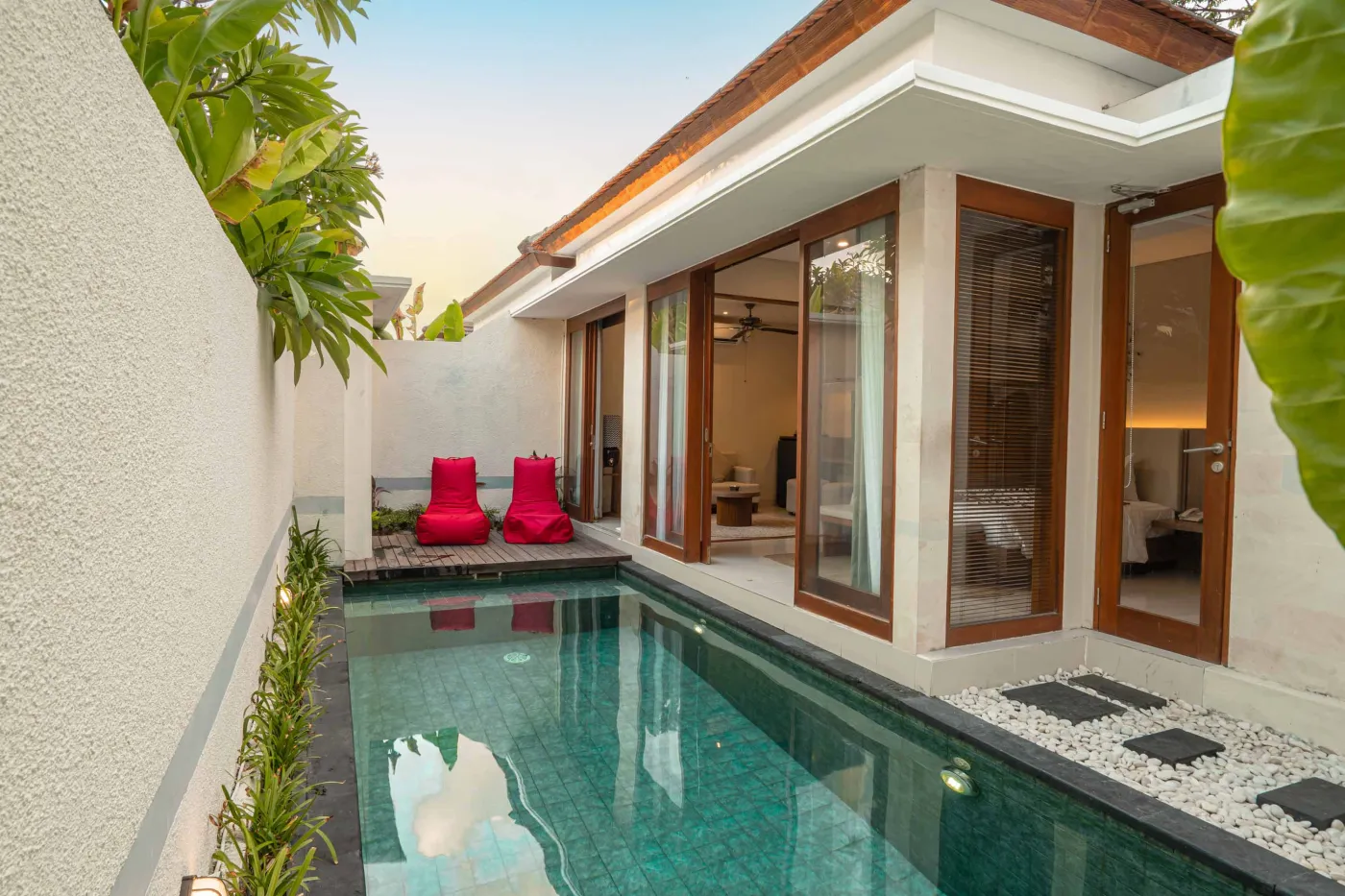 Modern Balinese villa with private infinity pool, red lounge chairs, open wooden doors, and lush tropical greenery at sunset. - Bali Villas