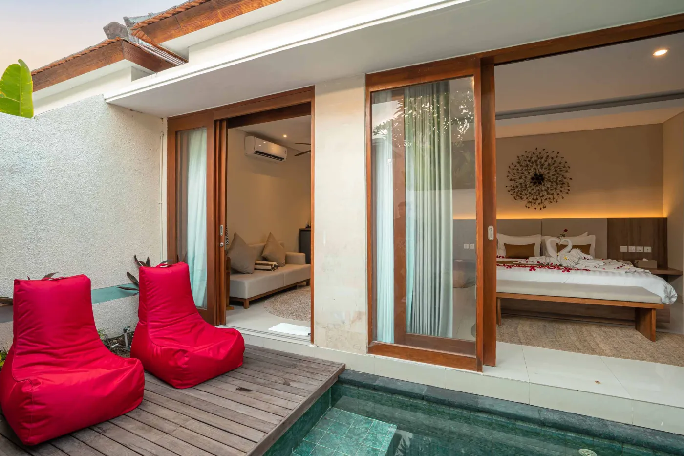 Luxurious Bali pool villa with private plunge pool, red bean bag chairs, and open doors to a rose petal-adorned bedroom. - Bali Villas