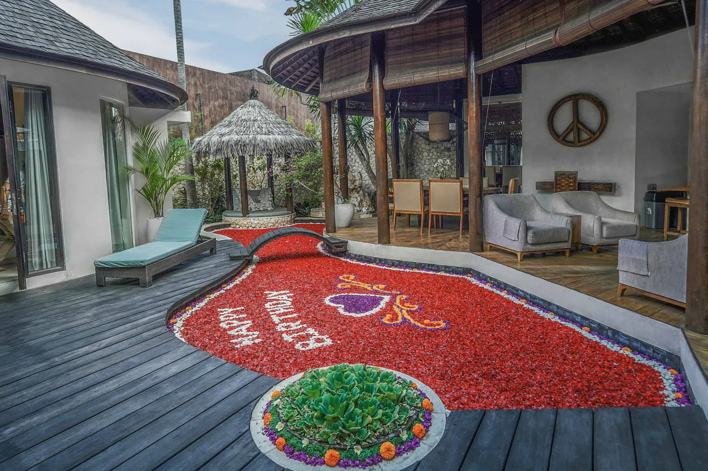 Modern open-plan kitchen with wooden island bar, four stools, stainless steel fridge, sink, and stove against a tropical stone wall with plants. - Bali Villas
