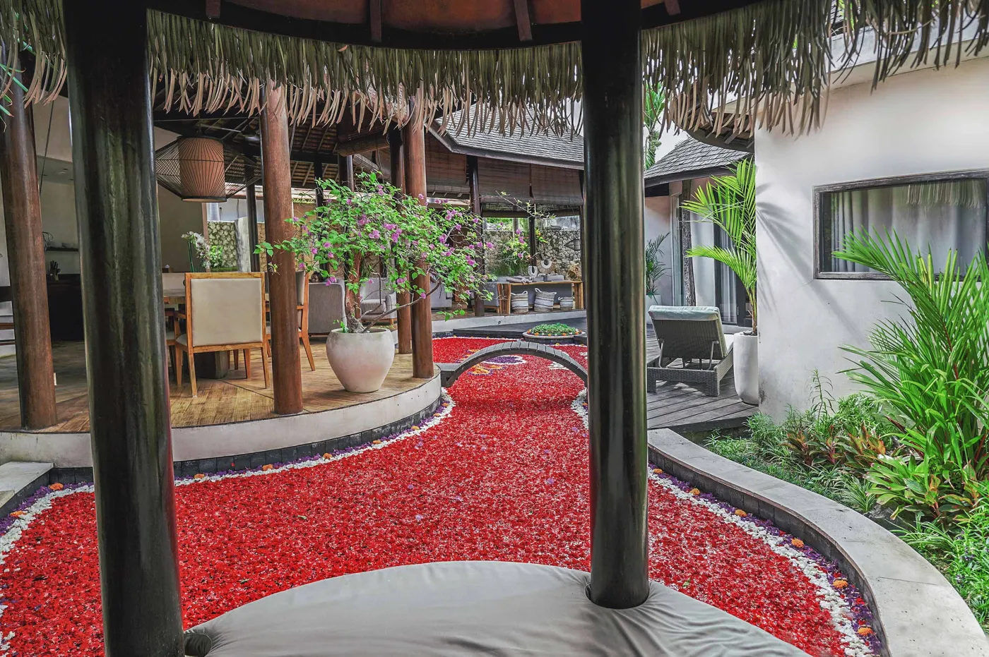 Modern open-plan kitchen with wooden island bar, four stools, stainless steel fridge, sink, and stove against a tropical stone wall with plants. - Bali Villas