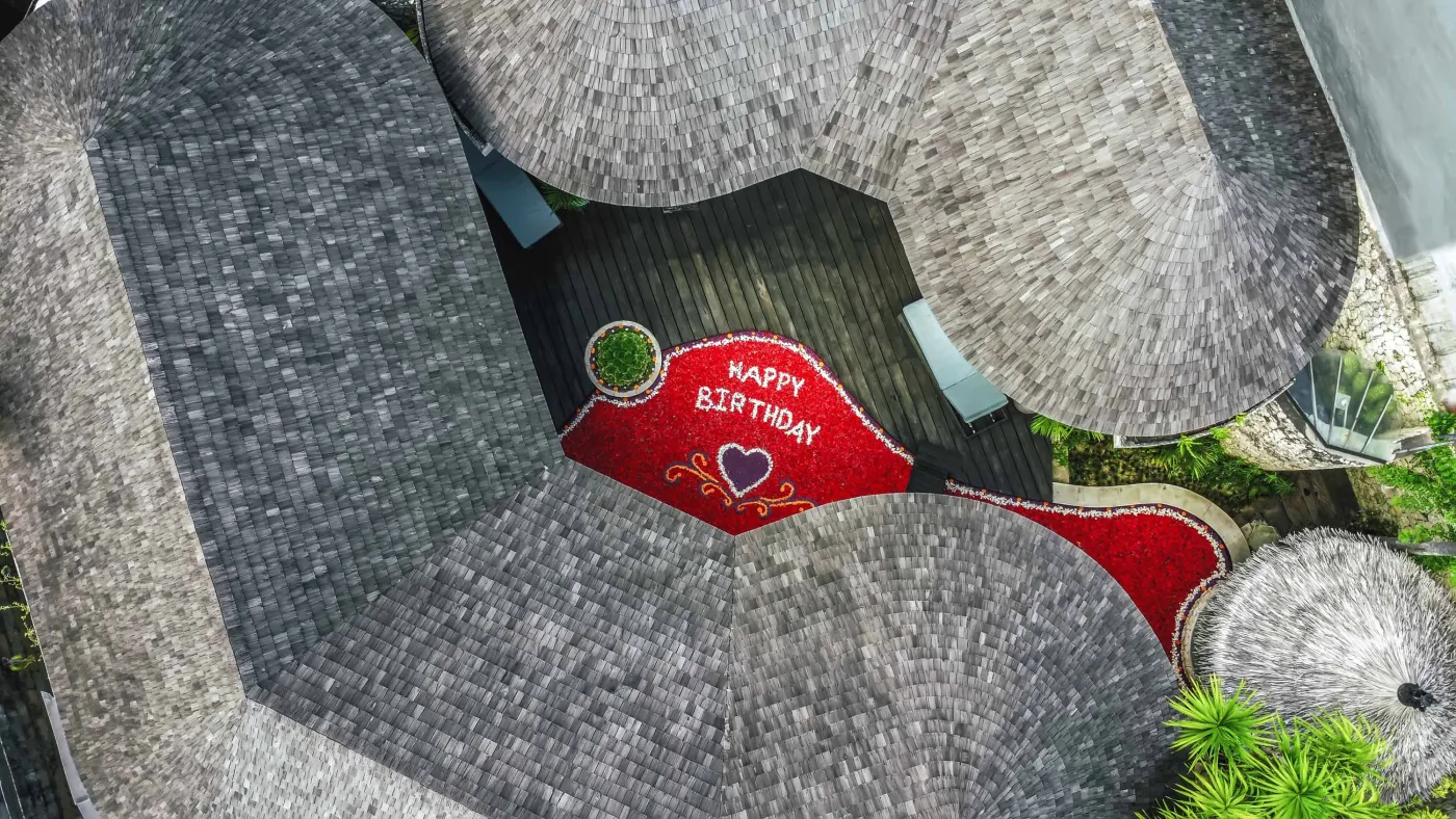 Modern open-plan kitchen with wooden island bar, four stools, stainless steel fridge, sink, and stove against a tropical stone wall with plants. - Bali Villas