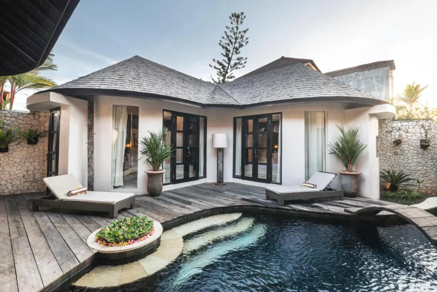 Modern open-plan kitchen with wooden island bar, four stools, stainless steel fridge, sink, and stove against a tropical stone wall with plants. - Bali Villas