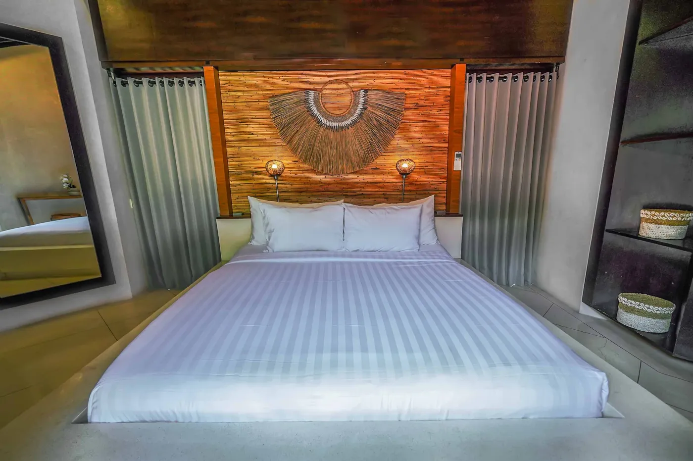 Modern open-plan kitchen with wooden island bar, four stools, stainless steel fridge, sink, and stove against a tropical stone wall with plants. - Bali Villas