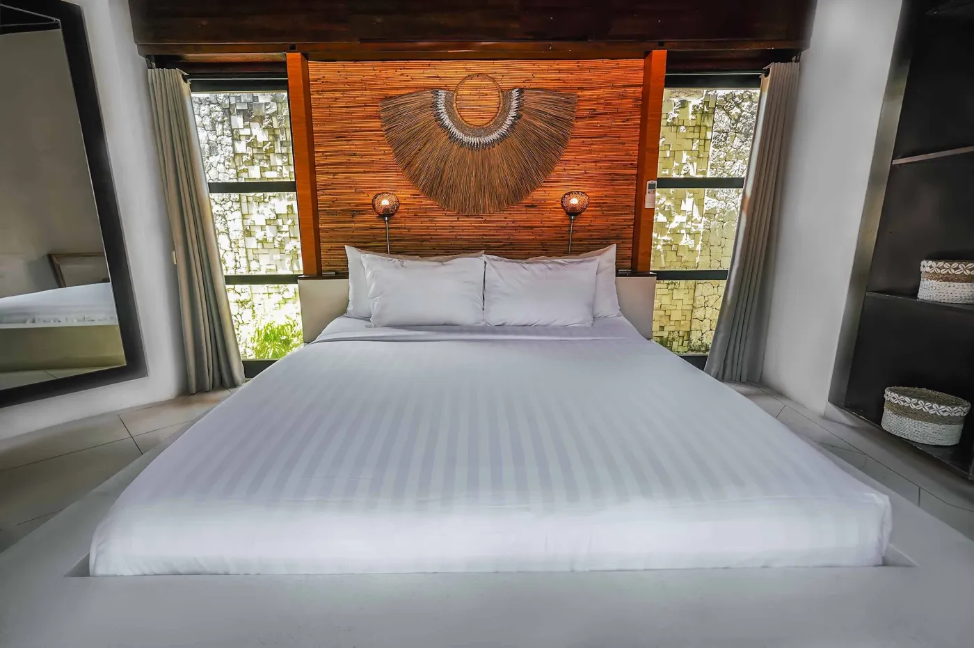 Modern open-plan kitchen with wooden island bar, four stools, stainless steel fridge, sink, and stove against a tropical stone wall with plants. - Bali Villas