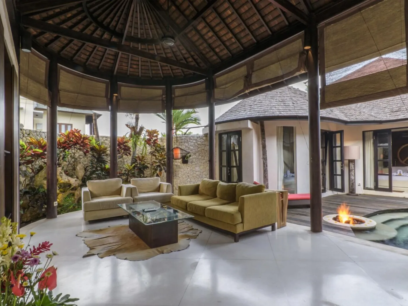 Modern open-plan kitchen with wooden island bar, four stools, stainless steel fridge, sink, and stove against a tropical stone wall with plants. - Bali Villas