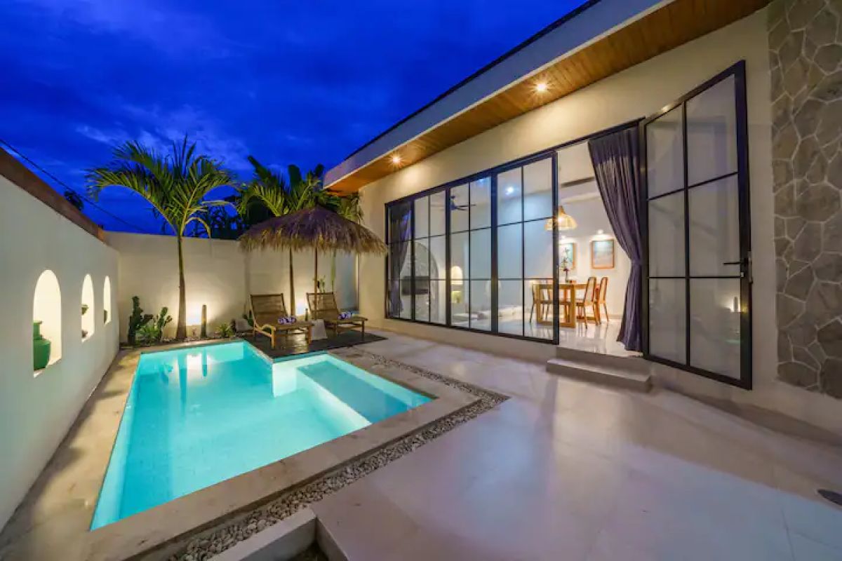 Modern kitchen and dining area in a tropical home, featuring rattan pendant lights, wooden table with chairs, and sliding glass doors to an outdoor pool at dusk. - Bali Villas