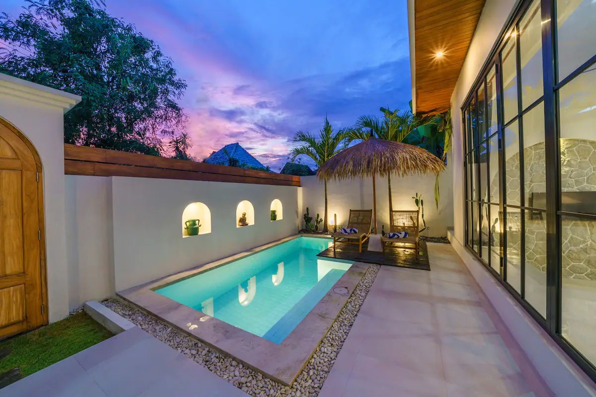 Modern kitchen and dining area in a tropical home, featuring rattan pendant lights, wooden table with chairs, and sliding glass doors to an outdoor pool at dusk. - Bali Villas