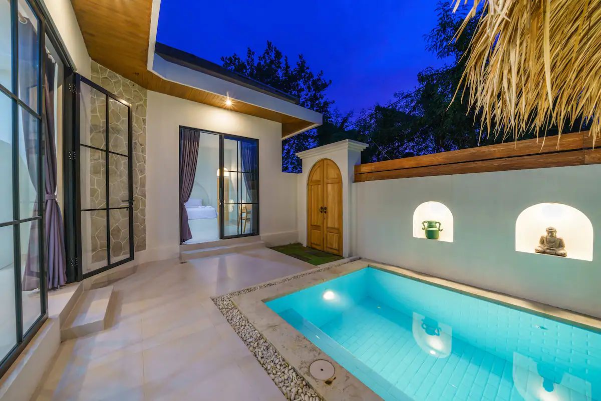Modern kitchen and dining area in a tropical home, featuring rattan pendant lights, wooden table with chairs, and sliding glass doors to an outdoor pool at dusk. - Bali Villas
