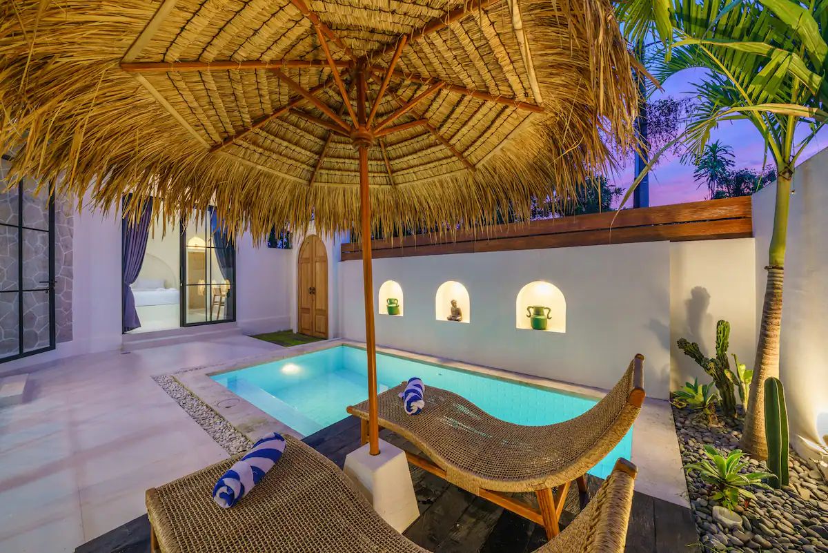 Modern kitchen and dining area in a tropical home, featuring rattan pendant lights, wooden table with chairs, and sliding glass doors to an outdoor pool at dusk. - Bali Villas