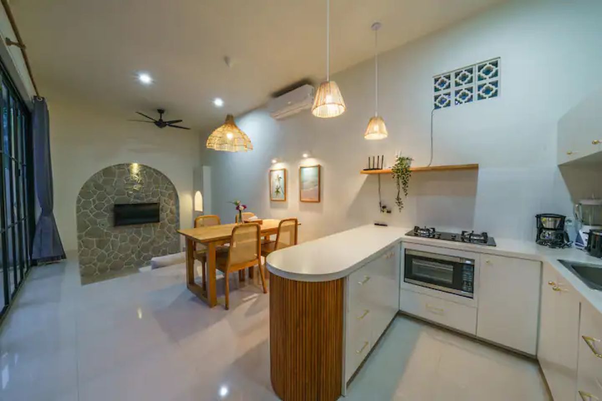 Modern kitchen and dining area in a tropical home, featuring rattan pendant lights, wooden table with chairs, and sliding glass doors to an outdoor pool at dusk. - Bali Villas
