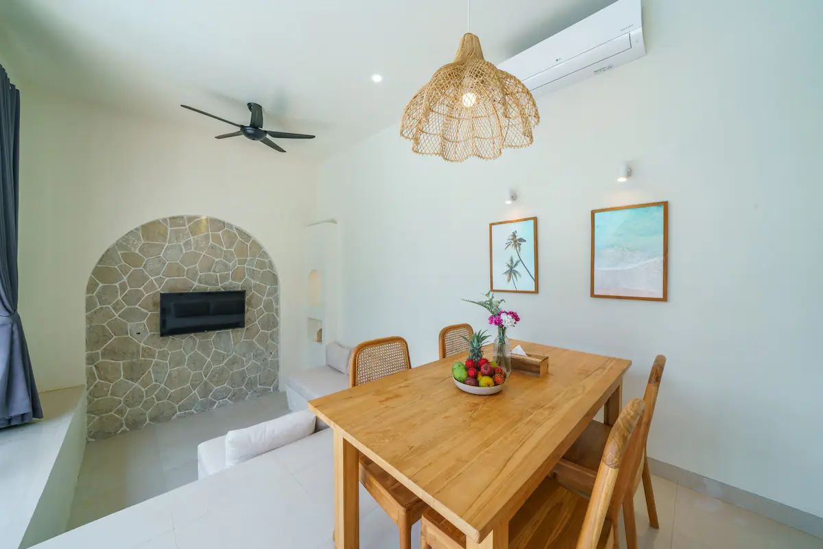 Modern kitchen and dining area in a tropical home, featuring rattan pendant lights, wooden table with chairs, and sliding glass doors to an outdoor pool at dusk. - Bali Villas