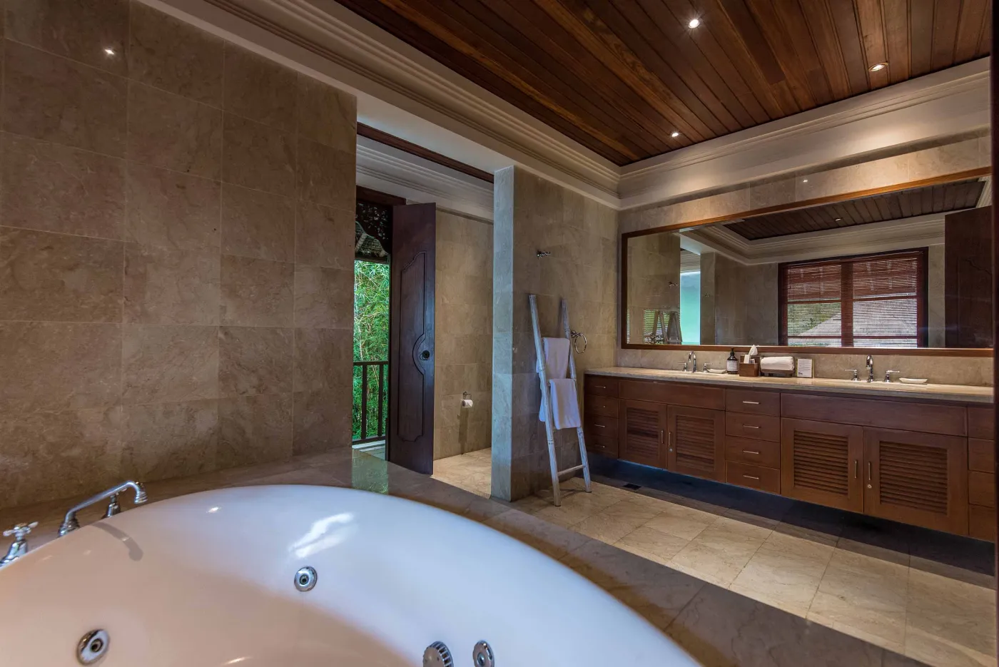 Luxurious tropical bathroom featuring a freestanding white bathtub, wooden vanity with double sinks, large mirror, and open door to lush greenery. - Bali Villas