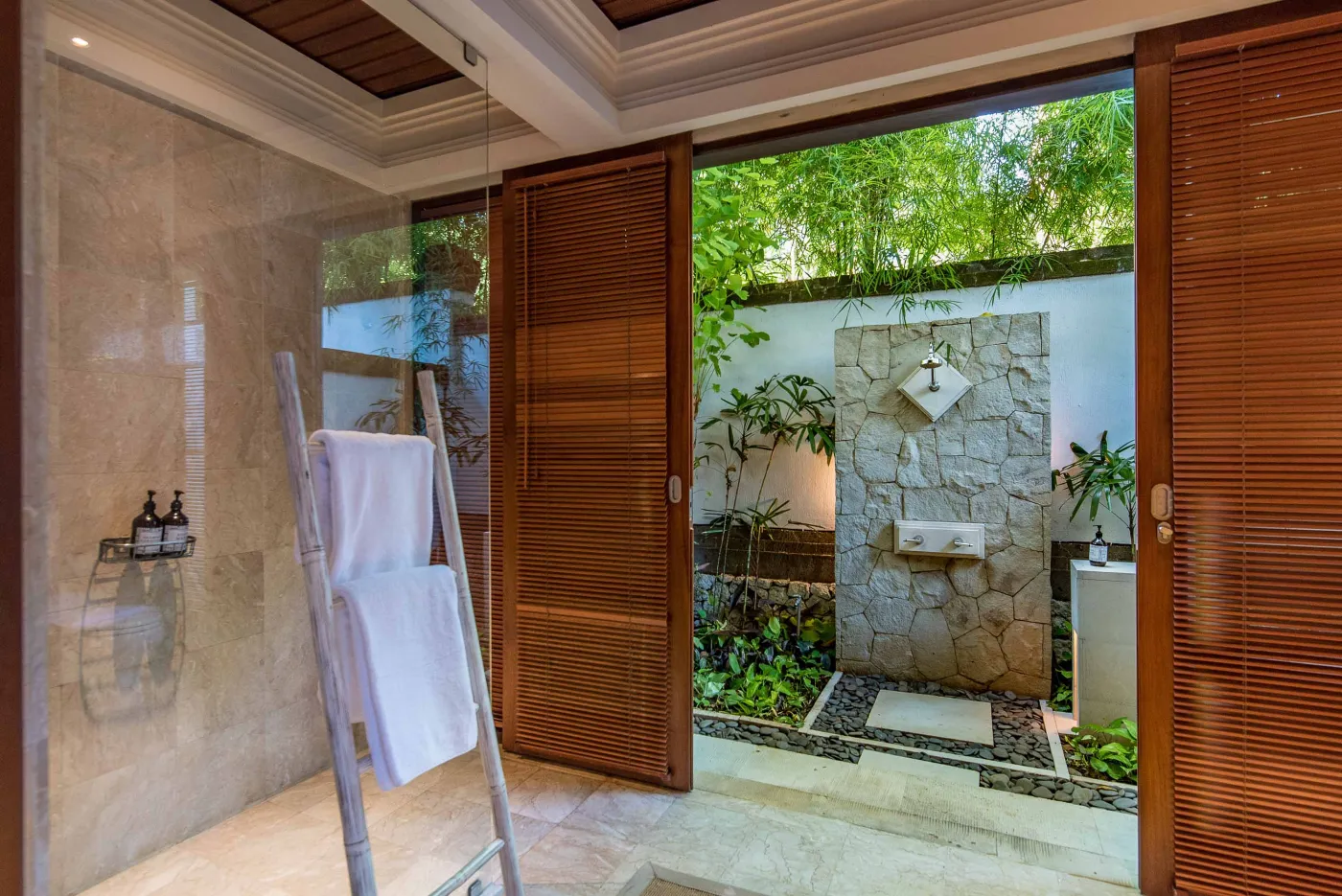 Luxurious tropical bathroom featuring a freestanding white bathtub, wooden vanity with double sinks, large mirror, and open door to lush greenery. - Bali Villas