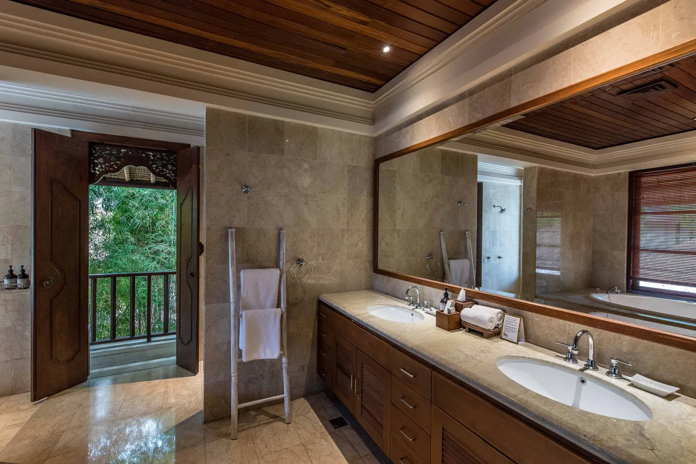 Luxurious tropical bathroom featuring a freestanding white bathtub, wooden vanity with double sinks, large mirror, and open door to lush greenery. - Bali Villas