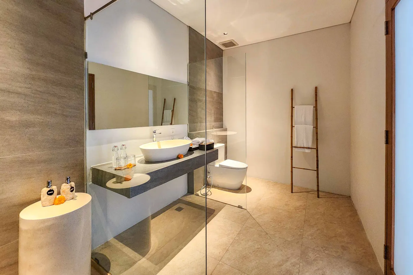 Modern luxury bathroom featuring a freestanding white bathtub, wooden vanity with towels and plants, and bamboo towel rack against neutral walls. - Bali Villas
