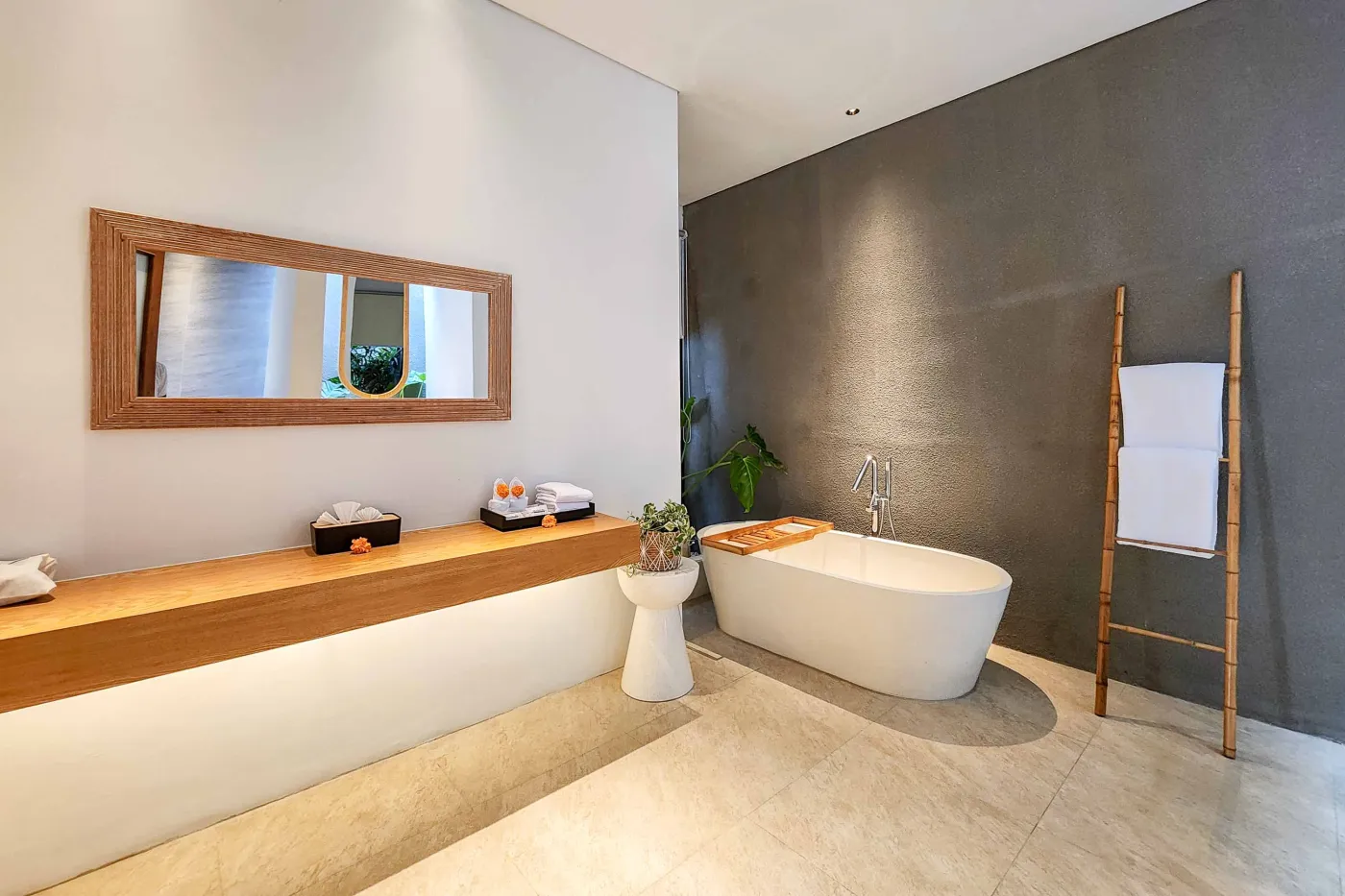 Modern luxury bathroom featuring a freestanding white bathtub, wooden vanity with towels and plants, and bamboo towel rack against neutral walls. - Bali Villas