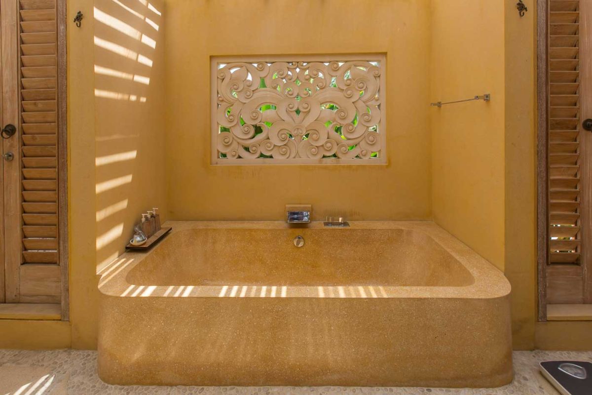 Serene open-air Balinese bathroom featuring a green freestanding bathtub with pink rose petals, tropical plants, and intricate carved wall panels. - Bali Villas