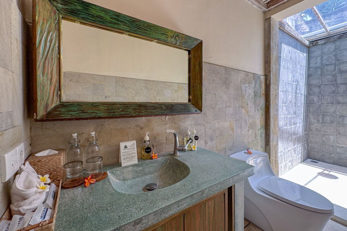 Serene open-air Balinese bathroom featuring a green freestanding bathtub with pink rose petals, tropical plants, and intricate carved wall panels. - Bali Villas