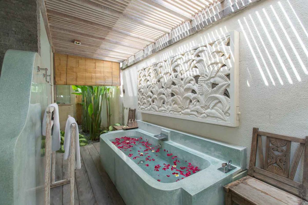 Serene open-air Balinese bathroom featuring a green freestanding bathtub with pink rose petals, tropical plants, and intricate carved wall panels. - Bali Villas