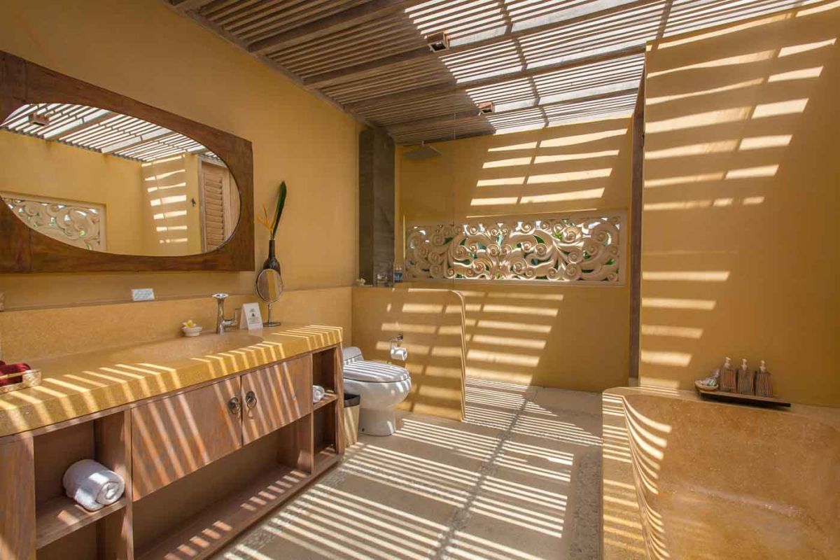 Serene open-air Balinese bathroom featuring a green freestanding bathtub with pink rose petals, tropical plants, and intricate carved wall panels. - Bali Villas