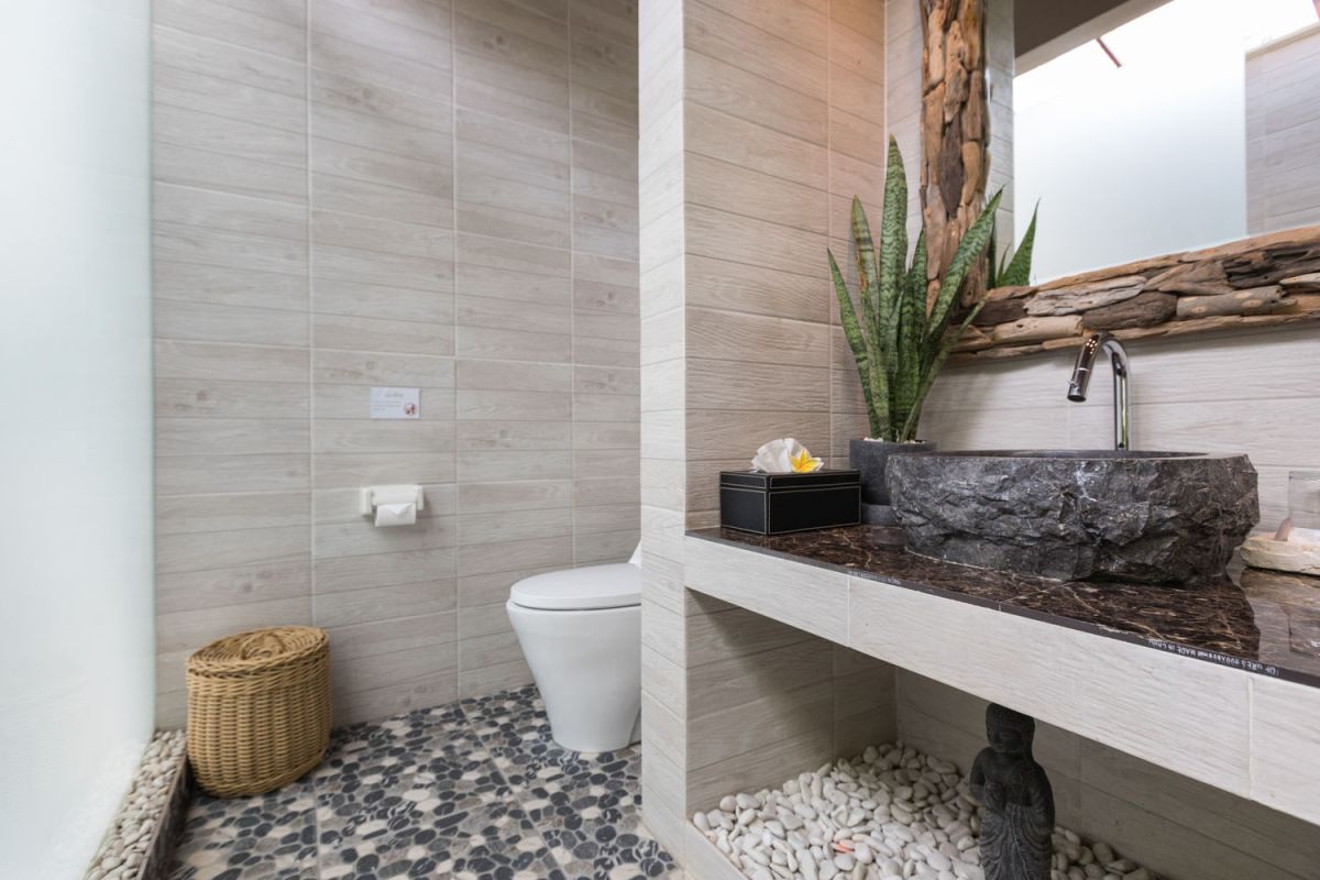 Modern Balinese bathroom with teak wood walls, pebble stone floor, rustic black stone sink, snake plants, and woven accents. - Bali Villas