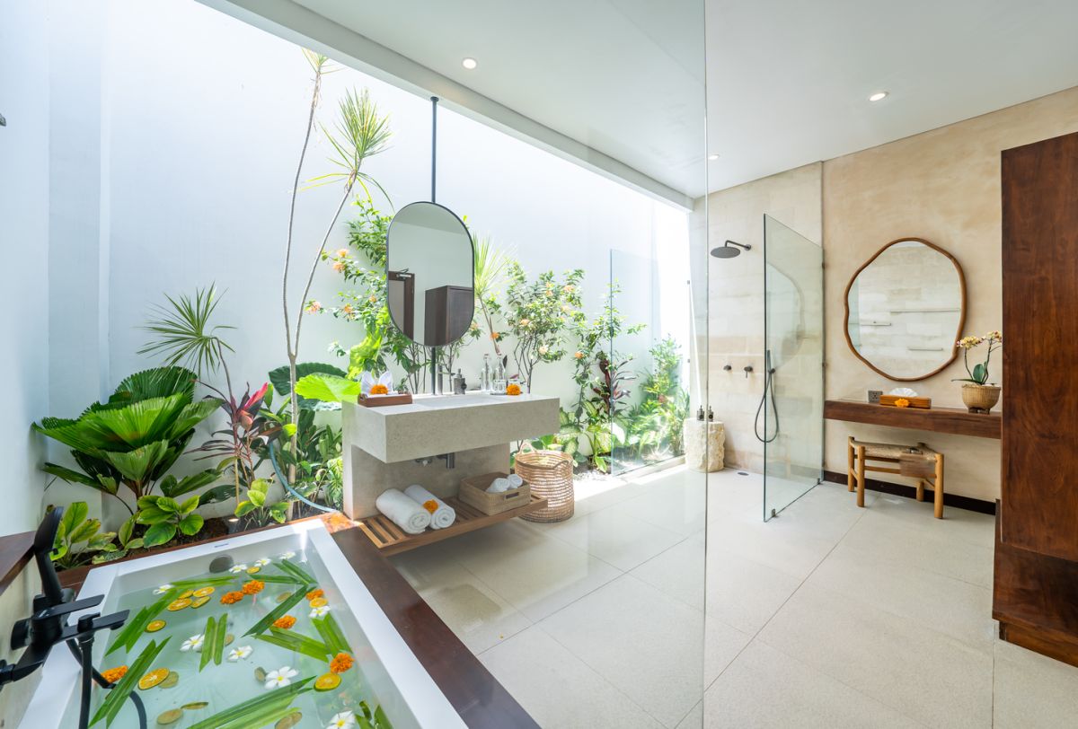 Modern luxury bathroom with soaking tub filled with water and lotus flowers, glass shower enclosure, tropical plants, and natural stone vanity. - Bali Villas