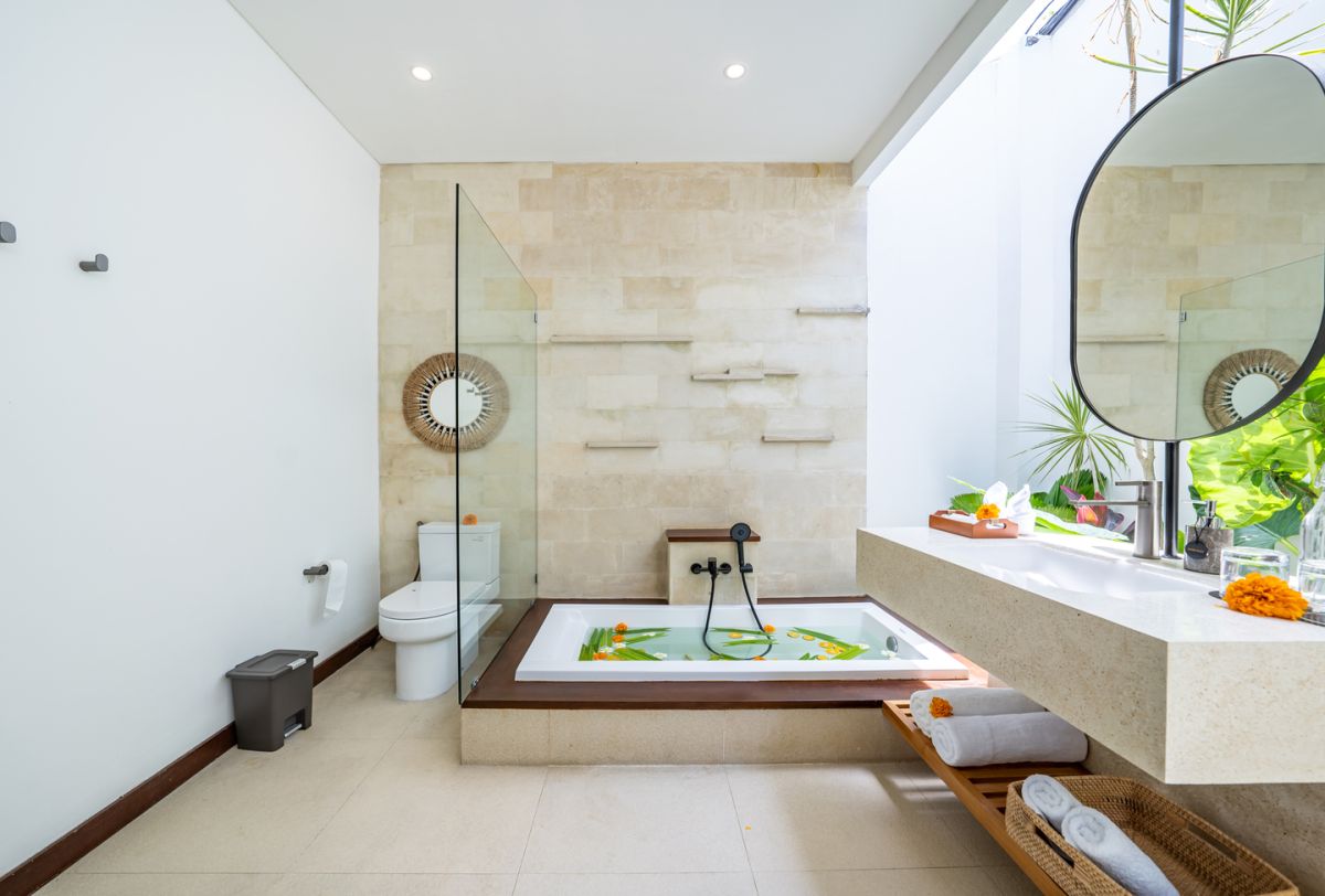 Modern luxury bathroom with soaking tub filled with water and lotus flowers, glass shower enclosure, tropical plants, and natural stone vanity. - Bali Villas