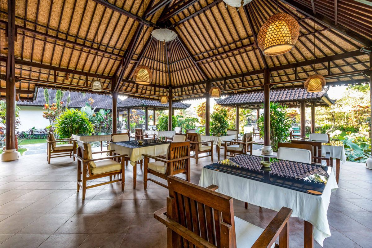 Spacious open-air Balinese pavilion restaurant with thatched roof, wooden tables set for dining, and lush tropical greenery. - Bali Villas