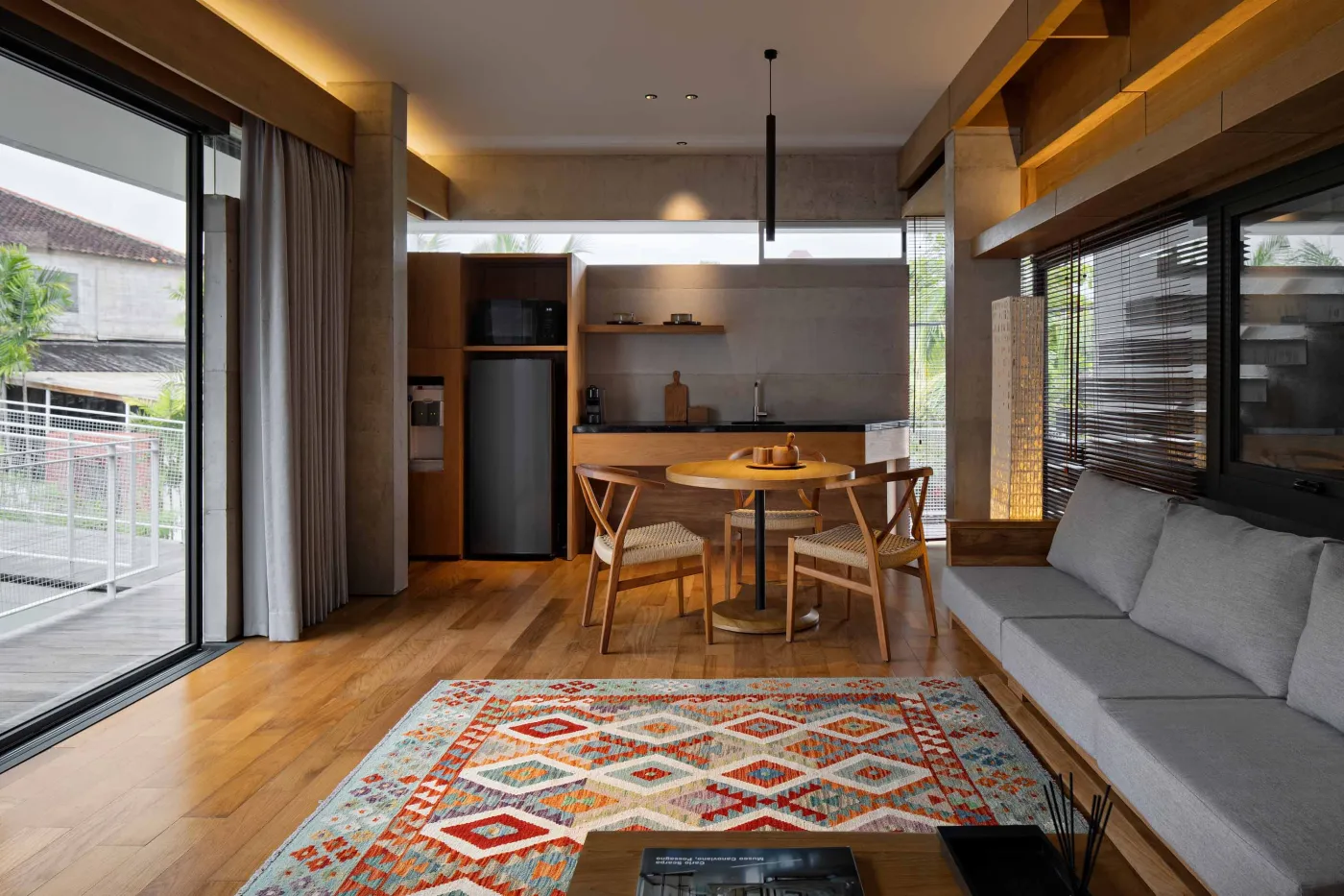 Modern wooden kitchen and dining area with pendant lights, long table set with bowl and runner, and large frosted windows overlooking greenery. - Bali Villas