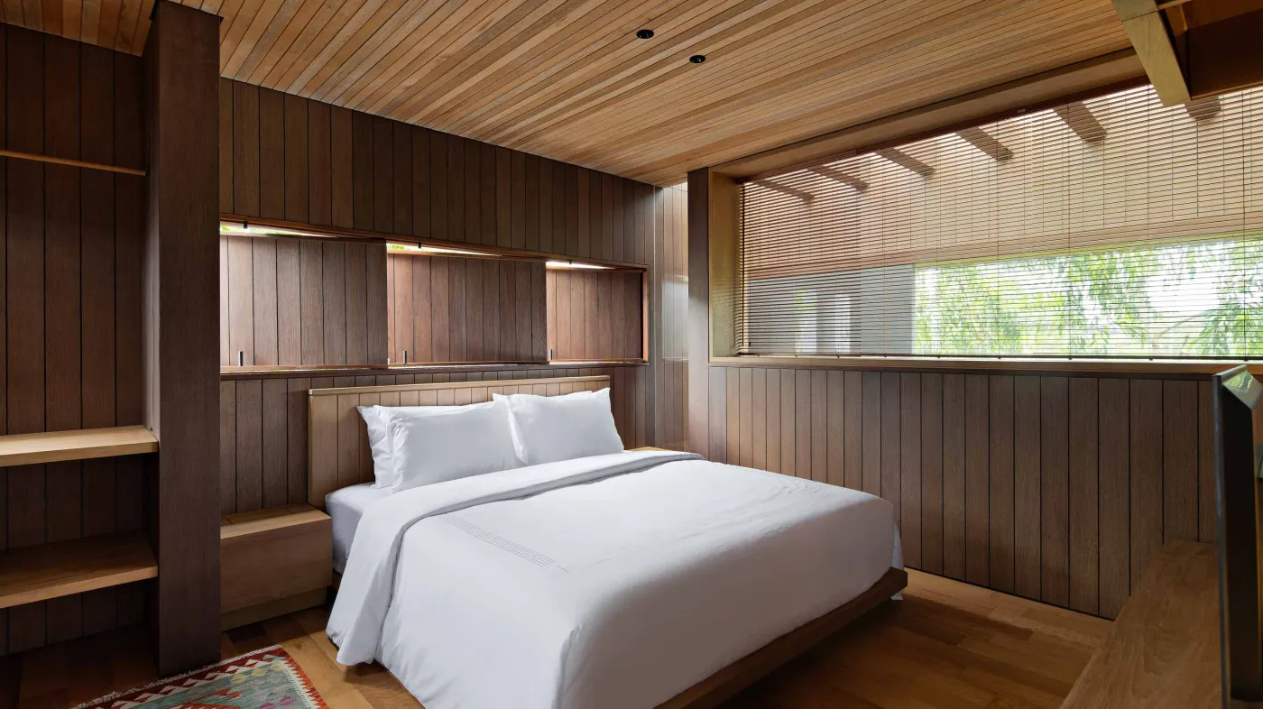 Modern wooden kitchen and dining area with pendant lights, long table set with bowl and runner, and large frosted windows overlooking greenery. - Bali Villas